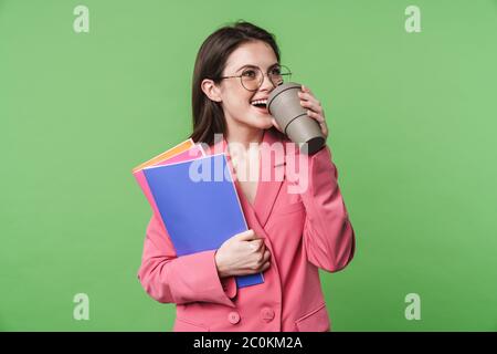Portrait of a beautiful happy young elegant woman wearing pink jacket standing isolated over green background, drinking takeaway coffee, holding folde Stock Photo