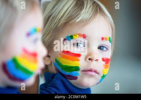 Beautiful blond toddler boy with rainbow painted on his face and messy hands, smiling happily Stock Photo