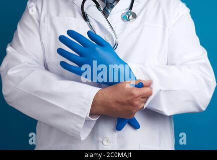 doctor in white uniform puts on his hands blue sterile latex gloves, blue background, close up Stock Photo