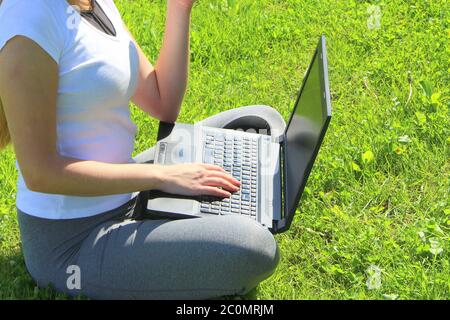 A beautiful young white girl in a white t-shirt and with long hair sitting on green grass, on the lawn and working behind a black laptop. Stock Photo