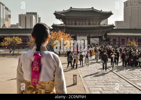 Girl in a traditional Korean costume hanbok observing tourists flowing through a gate into the Gyeongbokgung palace in Seoul South Korea Stock Photo
