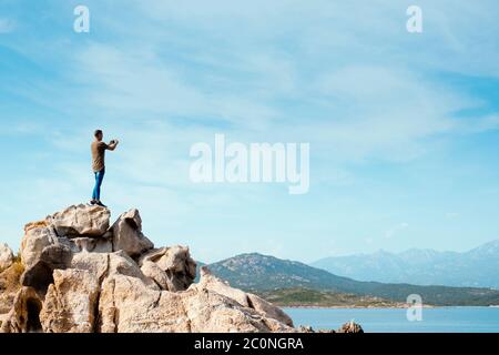 a young caucasian man, seen from behind, on the top of a rock formation, taking a photo of the sea in the Southern coast of Corsica, in France, with h Stock Photo