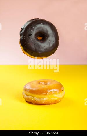 Chocolate doughnut falling on to plain glazed doughnut on vibrant pink and yellow background Stock Photo