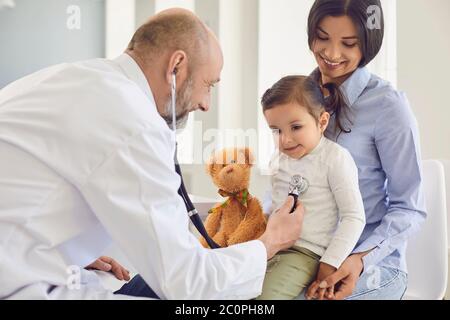 Family with a visit to the doctor in the clinic office. Senior doctor with a stethoscope listens to a little girl with mother patients in the hospital Stock Photo