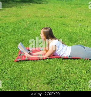 A beautiful young white girl in a white T-shirt and with long hair lies on a red plaid, on green grass, on the lawn and read book. Stock Photo