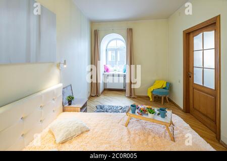Modern interior of luxury bedroom in apartment. Tray with breakfast on the bed. Arch window. Pillows on the sill. Wooden door. Stock Photo