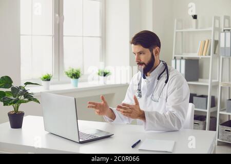 Male doctor providing medical consultation via video conference using laptop computer in office. Stock Photo