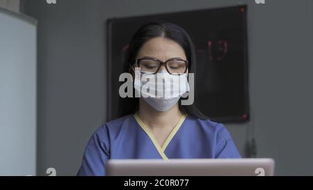 Doctor works on computer in office. Beautiful woman in mask, eyeglasses and blue uniform typing a laptop. Pandemic concept Stock Photo