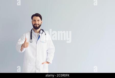 Male doctor in uniform showing thumb up gesture over grey background, copy space. Therapist approving medical service Stock Photo