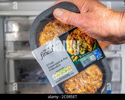 A man’s hand holding a pack of two Tesco Finest (own brand) cod, asparagus and pea fishcakes, coated in breadcrumbs, in front of a freezer. Stock Photo