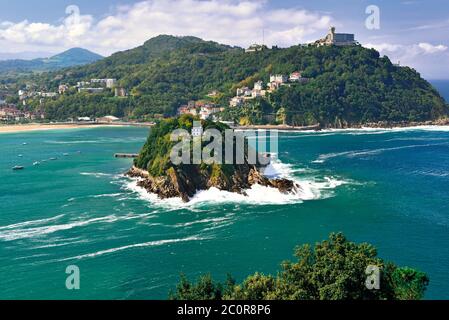 View to small green island in the middle of green ocean and hill with building at the top surrounded by sand beach (San Sebastian) Stock Photo
