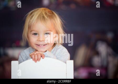 Beautiful blonde toddler boy, sitting on chair, playing with toys in a playroom Stock Photo