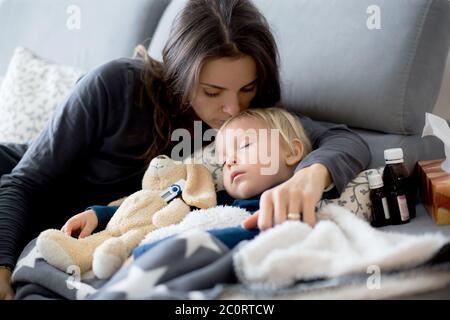 Blond toddler boy, sleeping on the couch in living room, lying down with fever, mom checking on him Stock Photo