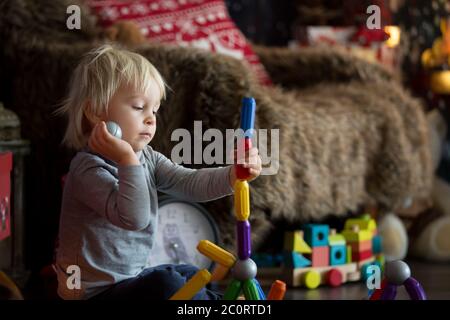 Sweet blonde toddler boy, playing with plastic construction, making different shapes, christmas decoration around him Stock Photo