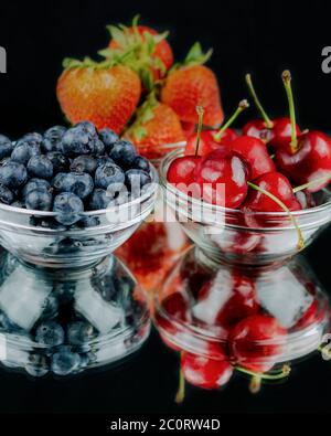 Three glass bowl filled with strawberries, cherries, and blueberries on a mirrow with reflection.  Black background. Stock Photo