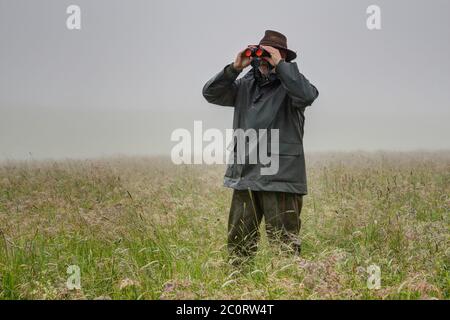 On a foggy, rainy day, a hunter stands in the high meadow and watches with his binoculars whether young deer are in the meadow. Stock Photo