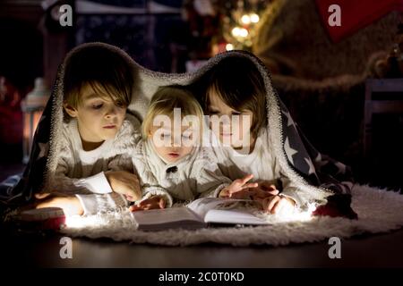 Three children, boy brothers, reading book at home at night on Christmas evening, lying on the floor Stock Photo