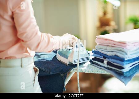 Closeup on woman in pink shirt and white pants with pile of folded ironed clothes ironing on ironing board in the house in sunny day. Stock Photo