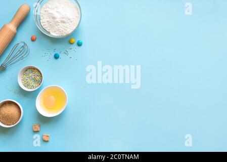 Baking utensils and ingredients.  Eggs, flour, sugar sprinkling, whisk and rolling pin on blue background. Baking concept flat lay, copy space. Stock Photo