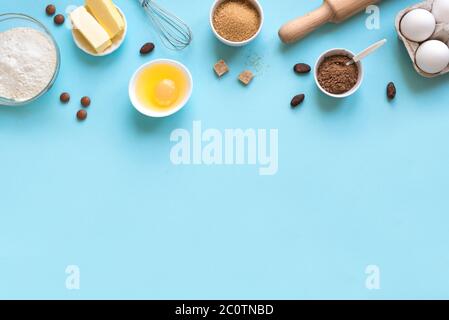 Baking utensils and ingredients.  Eggs, flour, cocoa beans and powder, sugar, whisk and rolling pin on blue background. Baking concept flat lay, copy Stock Photo