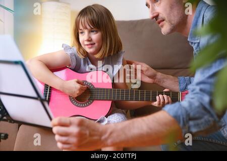 Little girl learning to play the guitar at home with a teacher in private lessons sitting on the living room sofa close up Stock Photo