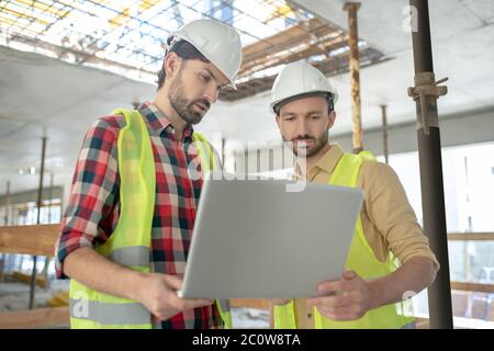 Building workers in yellow vests holding laptop, checking something on it Stock Photo