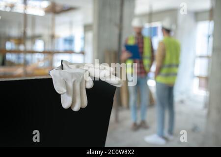 Close-up of protective gloves, building workers in yellow vests standing in the background, discussing something Stock Photo