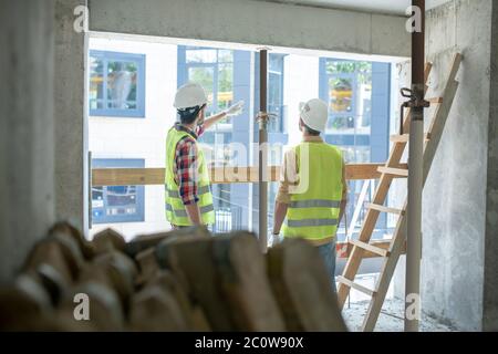 Building workers in yellow vests and gloves standing back to camera near the window, discussing, pointing at something Stock Photo