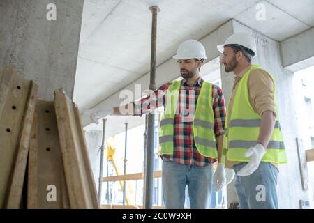 Building workers in yellow vests and gloves standing near pile of boards, pointing at them Stock Photo