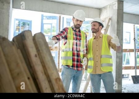 Building workers in yellow vests and gloves standing near pile of boards, discussing what to do with them, smiling Stock Photo
