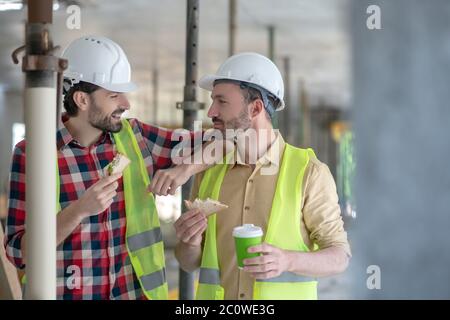 Building workers in yellow vests and helmets having lunch, talking smiling Stock Photo