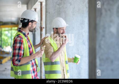 Building workers in yellow vests and helmets having coffee with sandwiches, discussing something Stock Photo