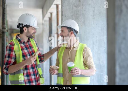 Building workers in yellow vests and helmets having coffee with sandwiches, talking, smiling Stock Photo