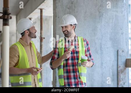 Building workers in yellow vests and helmets having coffee with sandwiches, discussing something, smiling Stock Photo