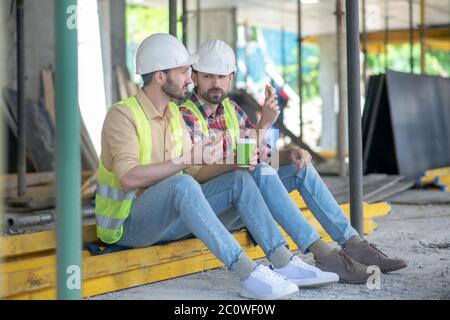 Building workers in yellow vests and helmets sitting on boards, having coffee with sandwiches, discussing something Stock Photo