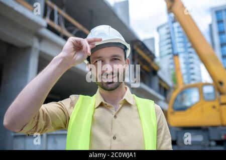 Building worker in helmet and yellow vest touching his helmet with his hand, smiling Stock Photo