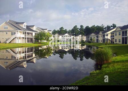 Background with a modern neighborhood with buildings around the pond. Houses and trees reflected in the tranquil water during beautiful cloudy morning Stock Photo
