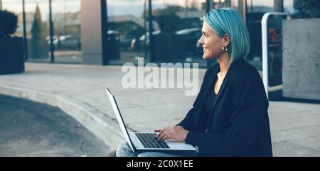 Young businesswoman with blue hair sitting in the street and working at the laptop Stock Photo