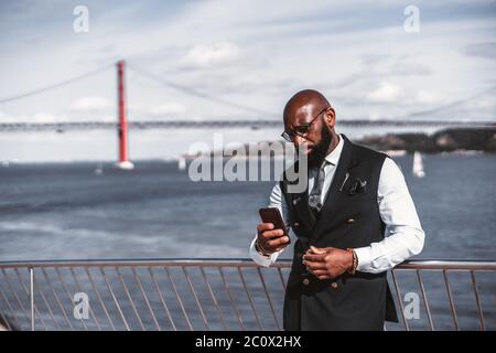 A serious elegant bald bearded African man entrepreneur in a costume and spectacles is using his smartphone while leaning against fencing with a river Stock Photo