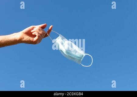 Man takes off medical protective mask and holds it on her finger on blue sky background, enjoys life, clean fresh air after Covid-19 pandemic, self-is Stock Photo