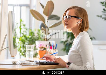 Pensive freelancer woman in glasses work on computer, using calculator to calculate the invoice, planning expenses from home office during self-isolat Stock Photo