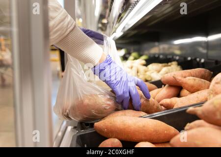 Woman wears rubber protective medical gloves, chooses sweet potato folding in disposable plastic bag in supermarket, close up. Protective measures aga Stock Photo