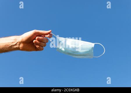 Man takes off medical protective mask and holds it on her finger on blue sky background, enjoys life, clean fresh air after Covid-19 pandemic, self-is Stock Photo