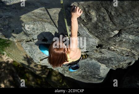 Horizontal snapshot of pretty woman in safety harness, climbing up the rock with rope, making next step helping with one hand. Top view. Concept of mountain sport, rock climbing Stock Photo