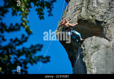 Brave lady alpinist in sportswear climbing extremely vertical rock under beautiful blue sky. Woman mountaineer ascending high mountain and trying to reach mountaintop. Concept of extreme sports. Stock Photo