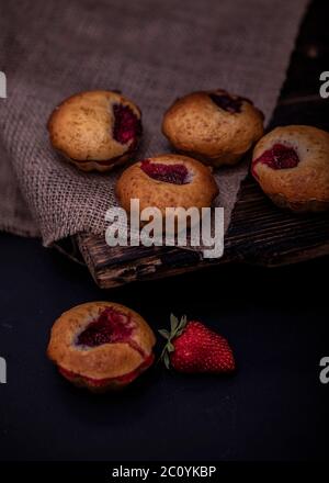 Cupcakes with white cream and strawberry on vintage blanket over black wooden background with fresh strawberries. Stock Photo