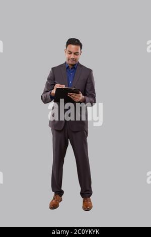 Businessman Writing in Clipboard Isolated. Indian Business man Standing Full Length with Clipboard in Hands Stock Photo