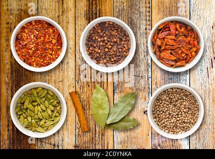 Spices including chilli flakes, cloves, bird's eye chillies, cardamoms, cinnamon stick, bay leaves and coriander seeds in white pots on a distressed w Stock Photo