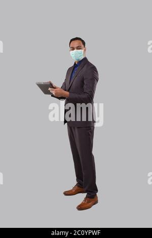 Businessman Using Tablet Wearing Medical Mask Isolated. Indian Business man Standing Full Length with Tablet in Hands Stock Photo