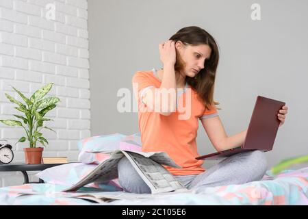 Girl sitting on the bed and laying a laptop on the bed looking through ads on a laptop screen Stock Photo